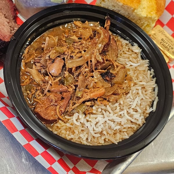 A black bowl of gumbo with rice, pulled pork, and vegetables, on a red and white checkered paper.
