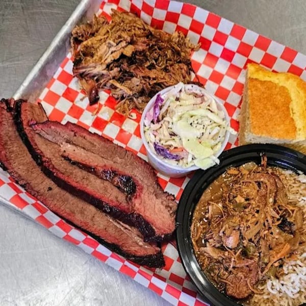 Barbecue platter with sliced brisket, pulled pork, coleslaw, cornbread, and gumbo on a red checkered tray.